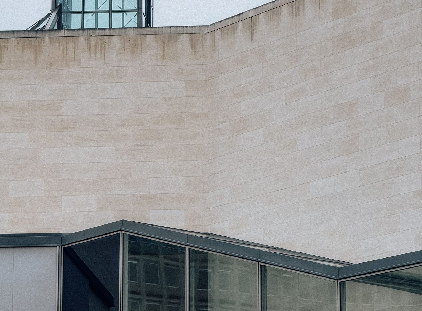 Pale limestone walls of a modern building meeting a dark-framed glazed roof